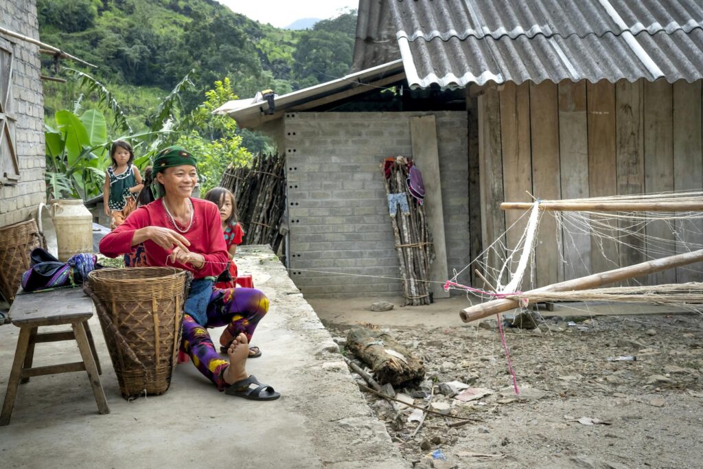 Smiling Vietnamese woman weaving outdoors in a rural village with children nearby.